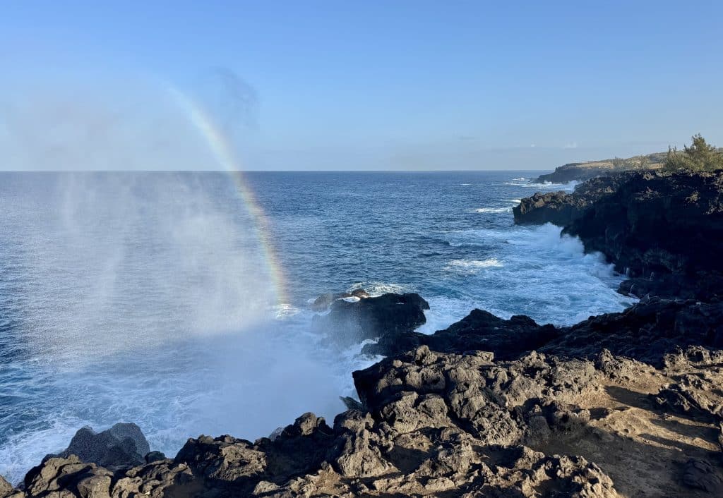 Angry waves crashing into black volcanic coastline, with mist and a rainbow.