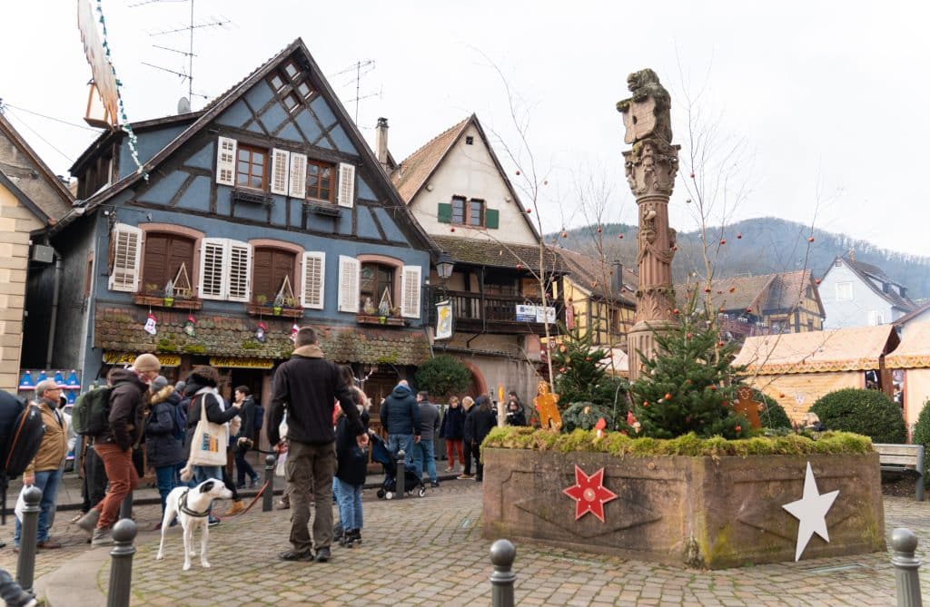 A small square in Ribeauville with a statue, and a half-timbered blue house with white shutters behind it. 