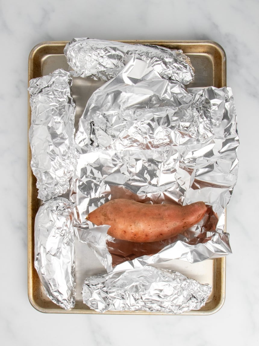 A metal baking sheet holding several sweet potatoes fully wrapped in aluminum foil, with one sweet potato uncovered in the center of the batch.
