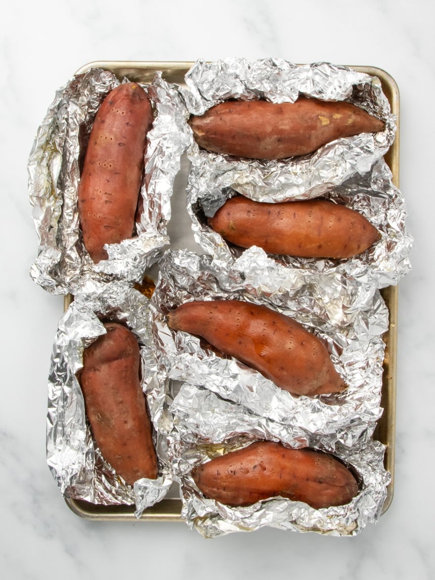 A metal baking sheet holding six cooked sweet potatoes, each resting in partially opened aluminum foil.