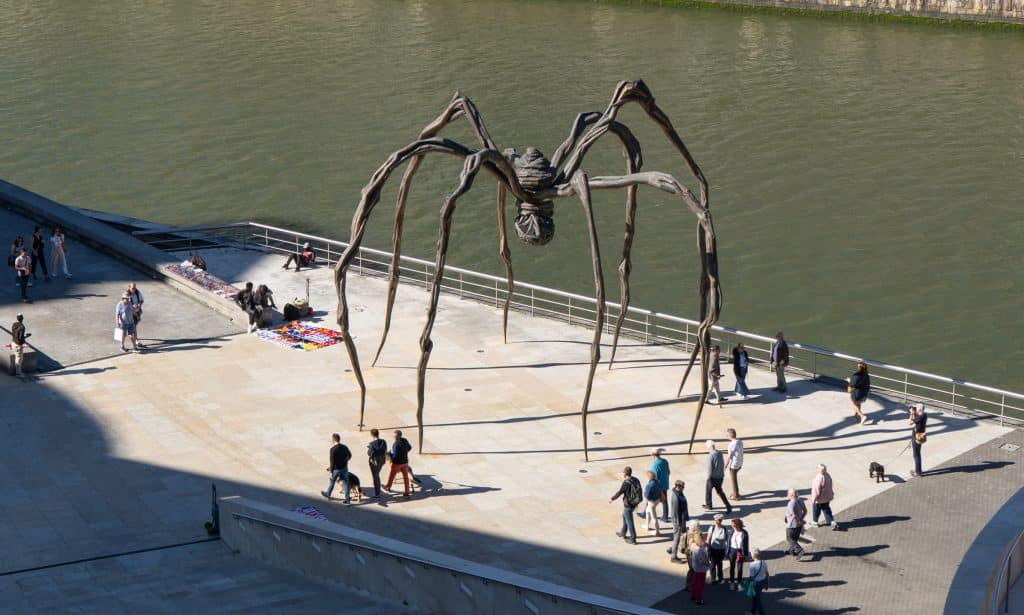 A giant spider sculpture on the edge of the river in Bilbao, lots of tiny people walking beneath it.