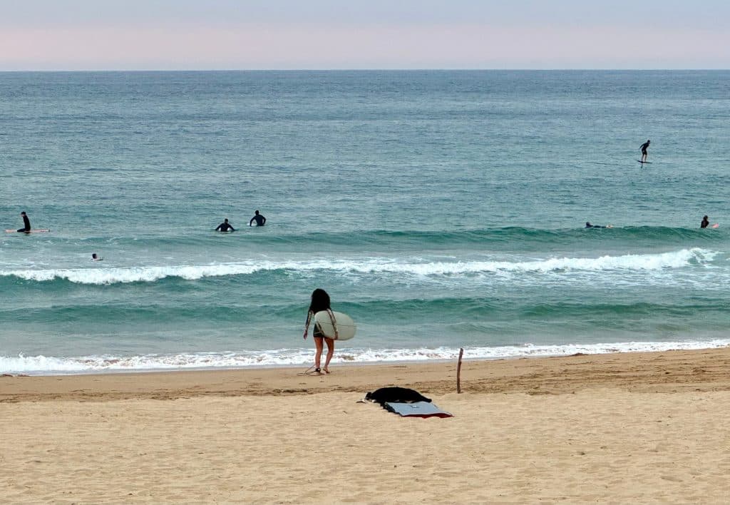 Woman running into the waves with a surfboard, other surfers in the water.