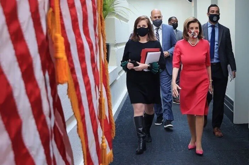 A group of people walk down a hallway lined with American flags. Terri McCullough, wearing a black dress and black mask, carries folders as she walks beside Nancy Pelosi, who is wearing a red dress and a patterned mask. Several masked staff members follow behind them.