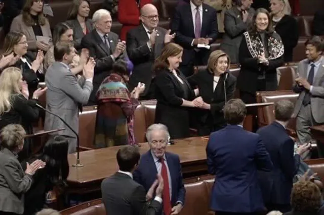 In the U.S. House chamber, Terri McCullough and Nancy Pelosi stand together near the front as people in the chamber applaud. McCullough and Pelosi hold hands while members seated and standing around them clap.