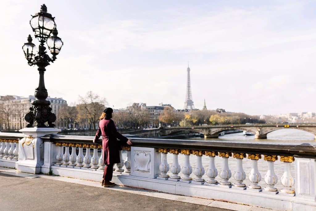 A woman standing on a bridge in Paris, looking at the Eiffel Tower in the distance. It's a cold, white-sky day and she wears a burgundy coat and hat.