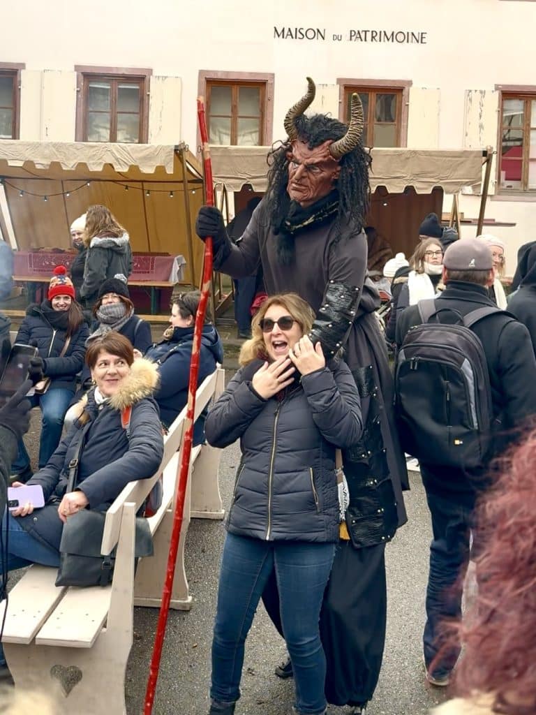A man on stilts in a devil mask and cloak grabs a woman around the neck as if to choke her. Her mouth is open in shock, but she's excited.