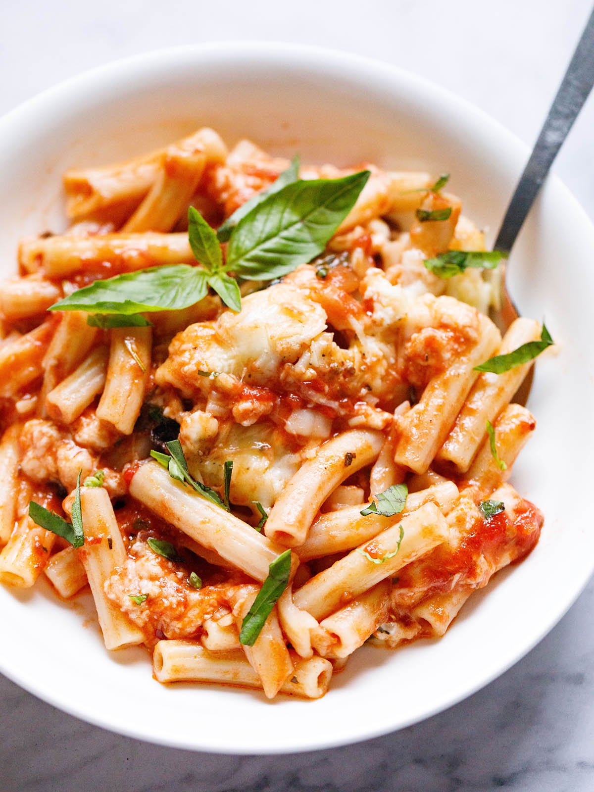 overhead shot of white bowl filled with a helping of three cheese vegetarian baked ziti and garnished with basil chiffonade and a silver fork