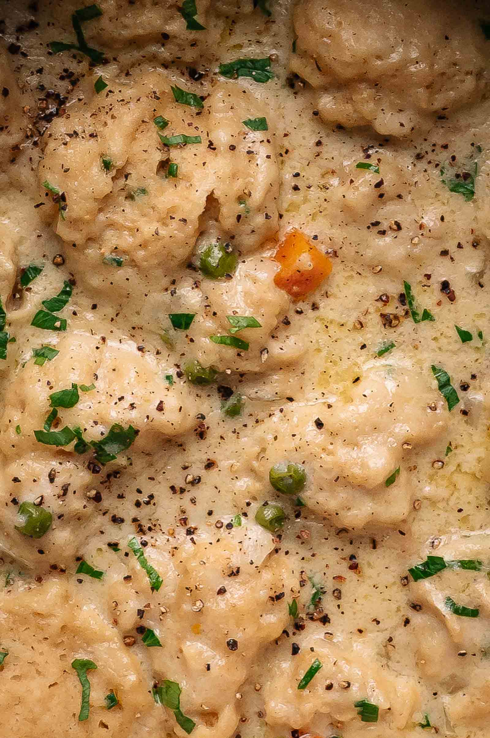 Creamy chicken stew with dumplings, peas, carrots, chopped herbs, and cracked black pepper visible on top.