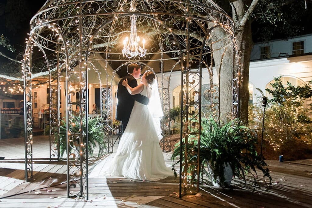 A couple dances under a chandelier inside a metal pergola outside in a courtyard