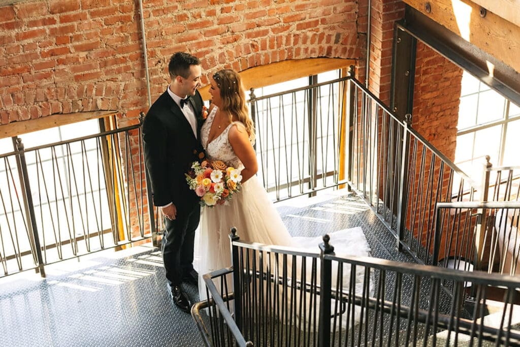 A bride and groom steal a private moment in a stairwell next to huge windows in a rustic space with brick walls and wooden beams