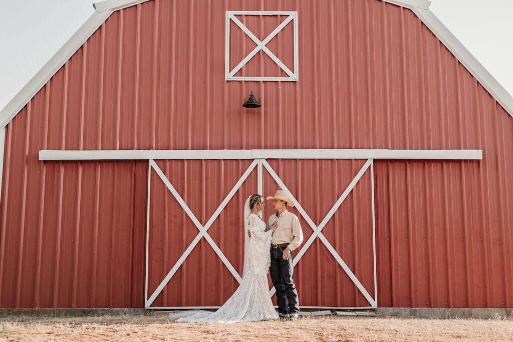 A bride and groom stand side by side in front of a bright red barn