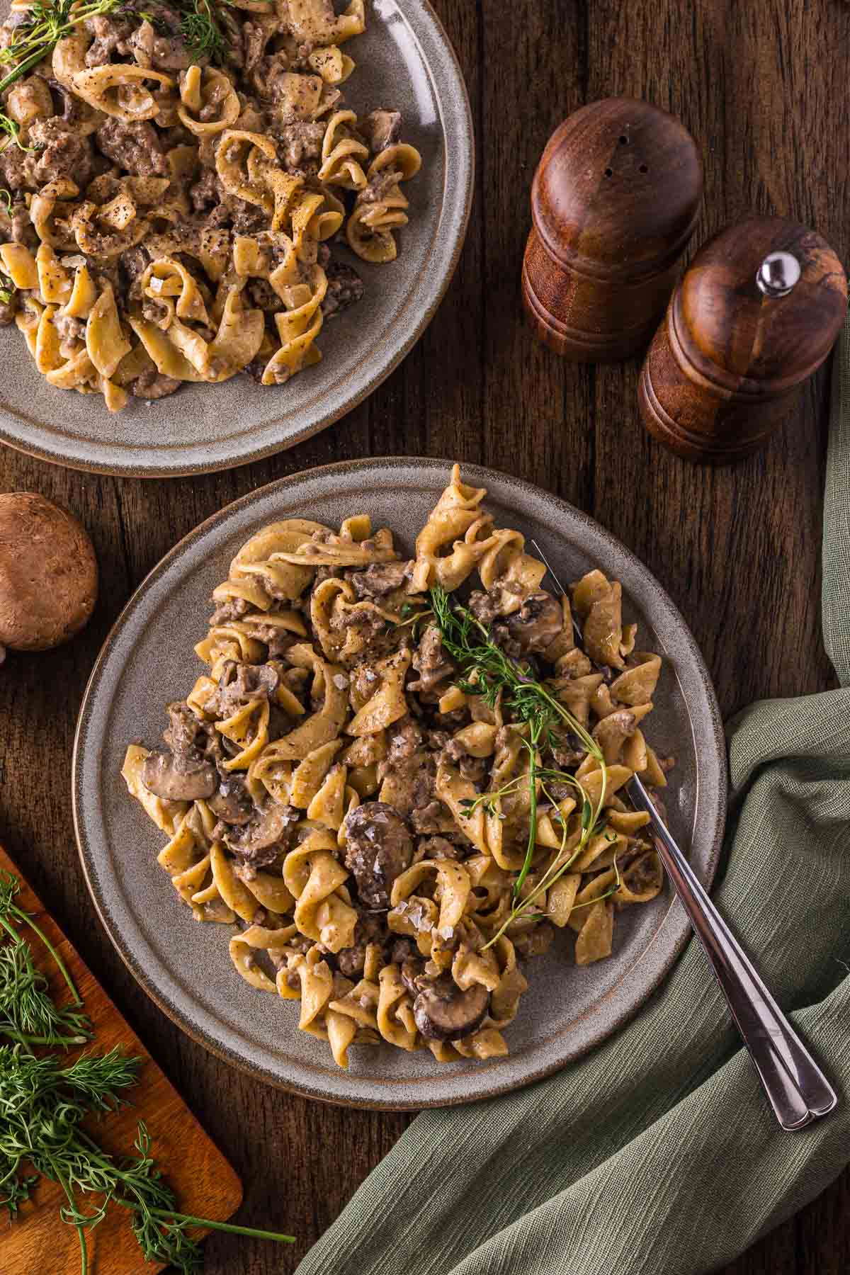 Two plates of egg noodles with a creamy mushroom and ground beef stroganoff, garnished with fresh dill, are set on a wooden table next to a green napkin, wooden salt and pepper shakers, and mushrooms.