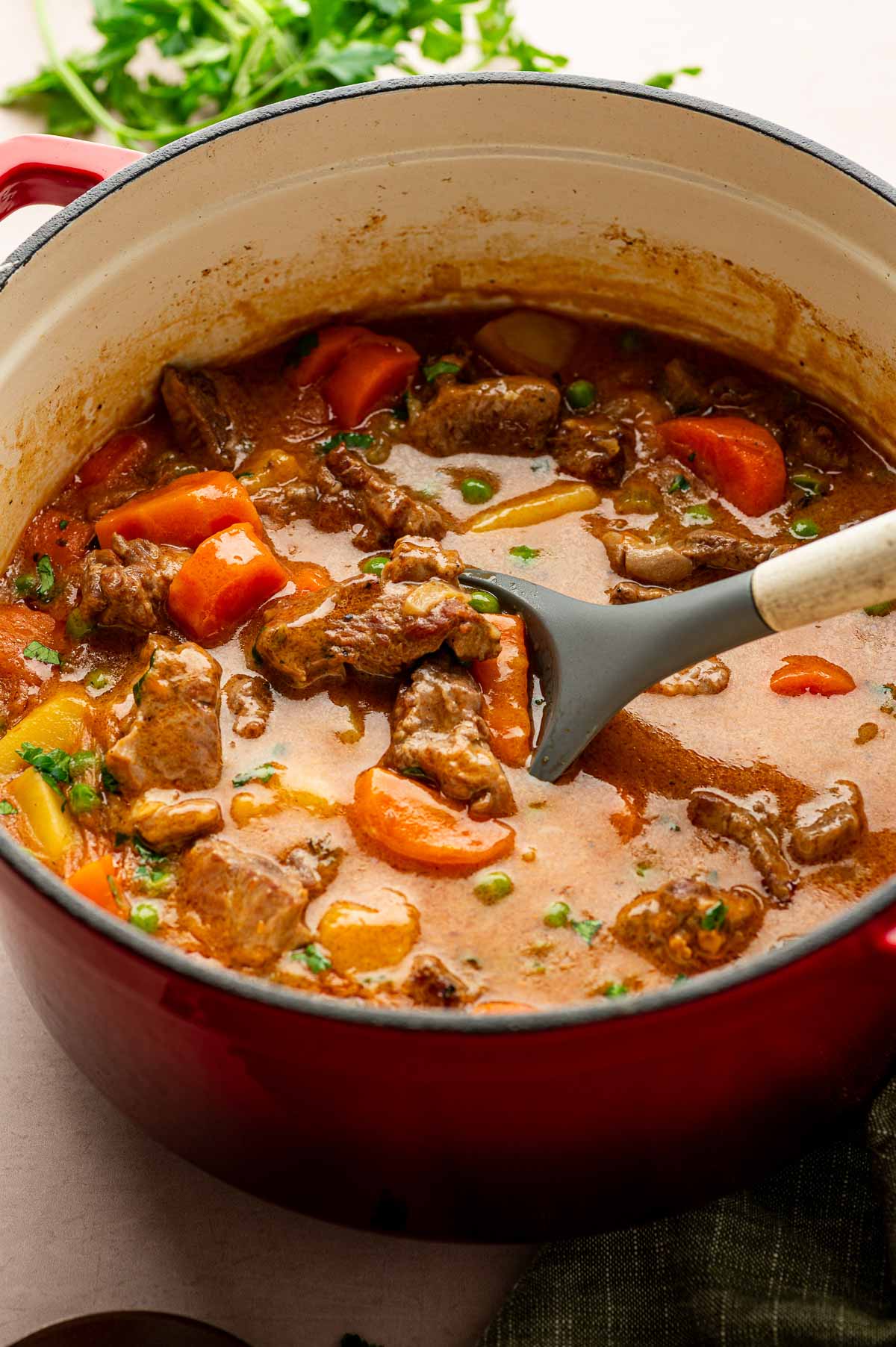 A red Dutch Oven filled with lamb stew containing chunks of cubed lamb, carrots, peas, and a brown broth. A ladle is resting inside the pot.