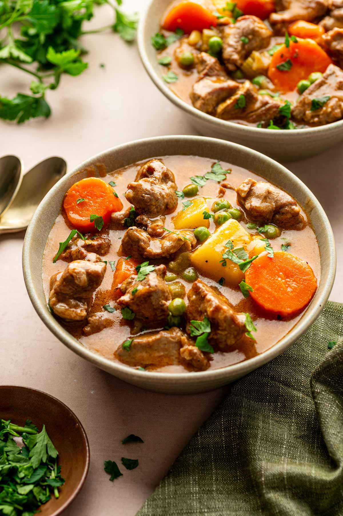 A bowl of lamb stew with chunks of lamb shoulder, carrots, potatoes, peas, and chopped parsley garnish, placed on a table with utensils and fresh herbs nearby.