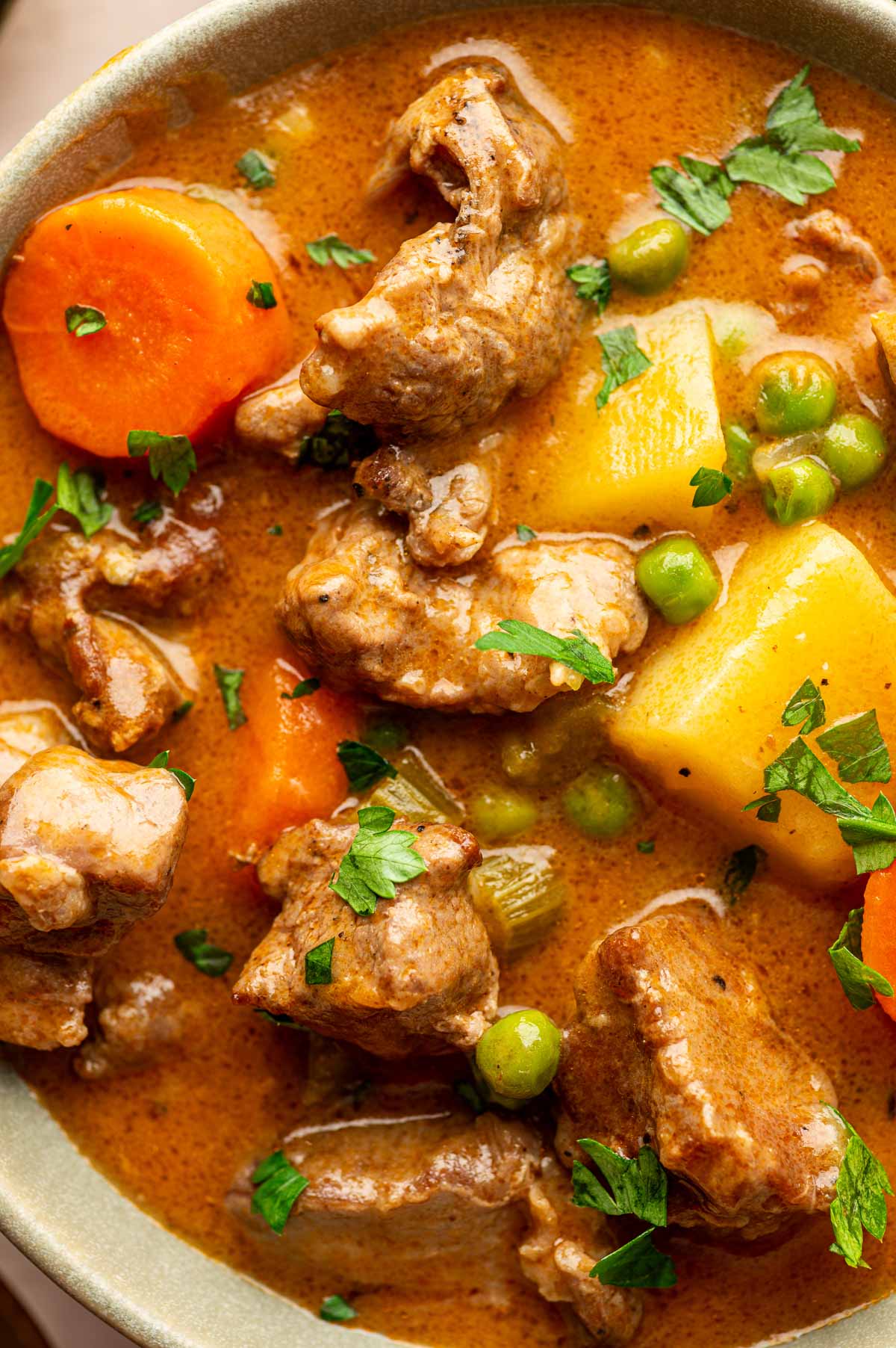 A close-up of lamb stew with chunks of lamb, potatoes, carrots, green peas, and fresh parsley in a brown broth.