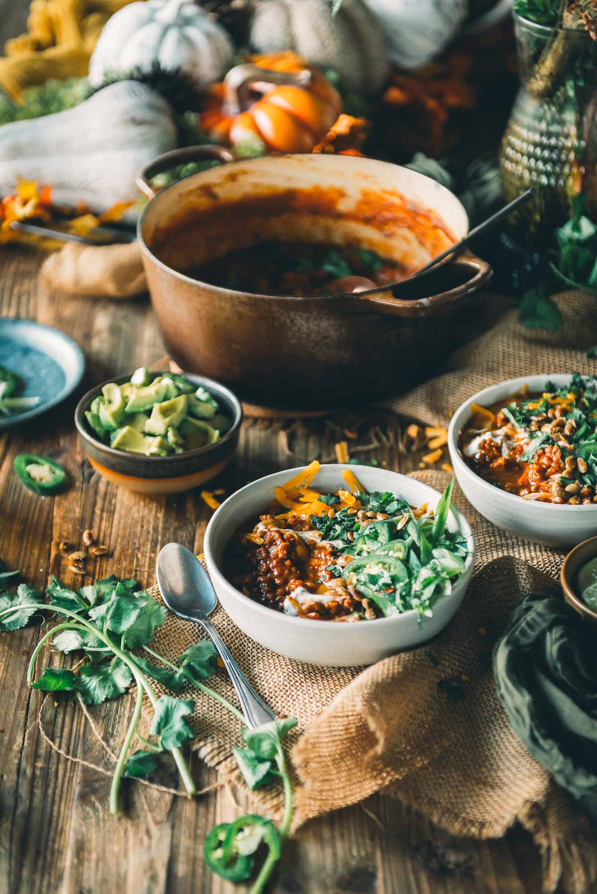 Two bowls of pumpkin chili with ground turkley topped with cheese and cilantro sit on a rustic wooden table, surrounded by a pot of chili, avocado slices, and scattered herbs and jalapeño slices.