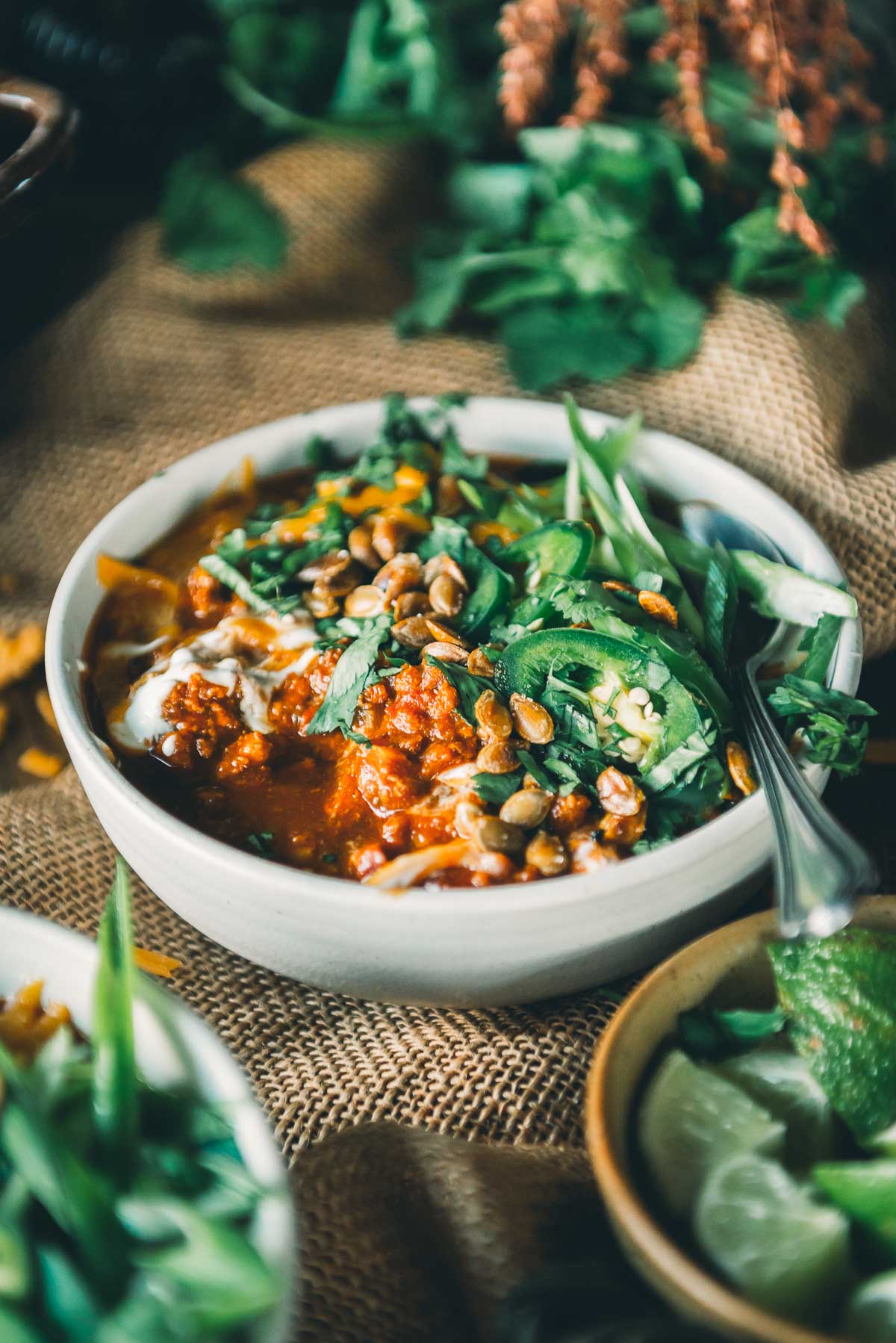 A bowl of pumpkin chili with ground turkley topped with greens, seeds, and herbs, placed on a burlap surface with lime wedges and more greens in the background.