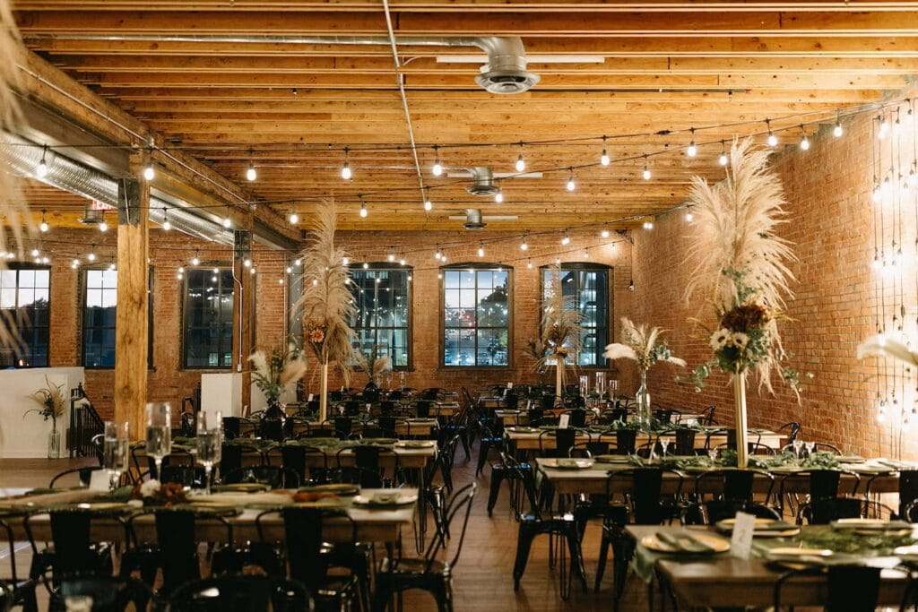 Wooden beams and brick walls set the tone for a rustic wedding reception in an industrial building hung with string lights and decorated with tall floral arrangements set atop long tables
