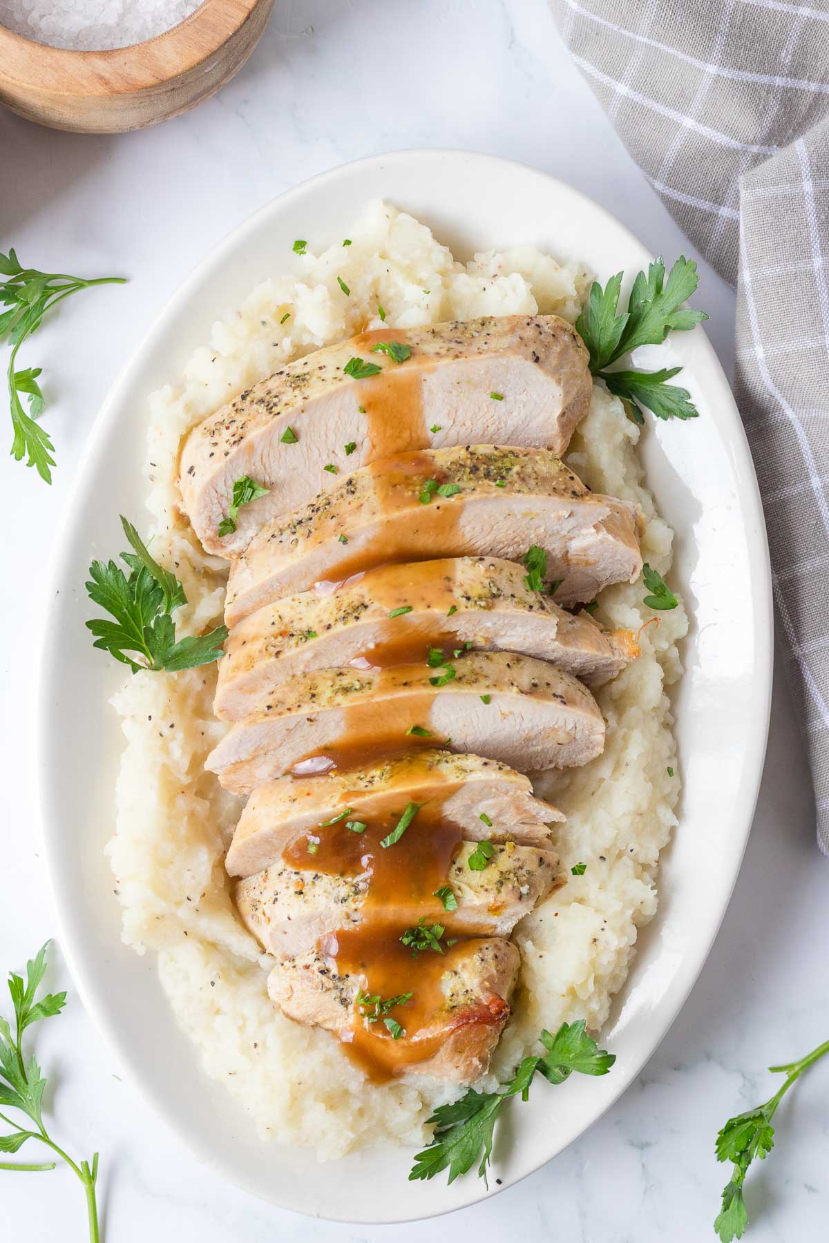 Sliced slow cooker turkey breast with brown gravy served on a bed of mashed potatoes, garnished with parsley on a white oval plate.