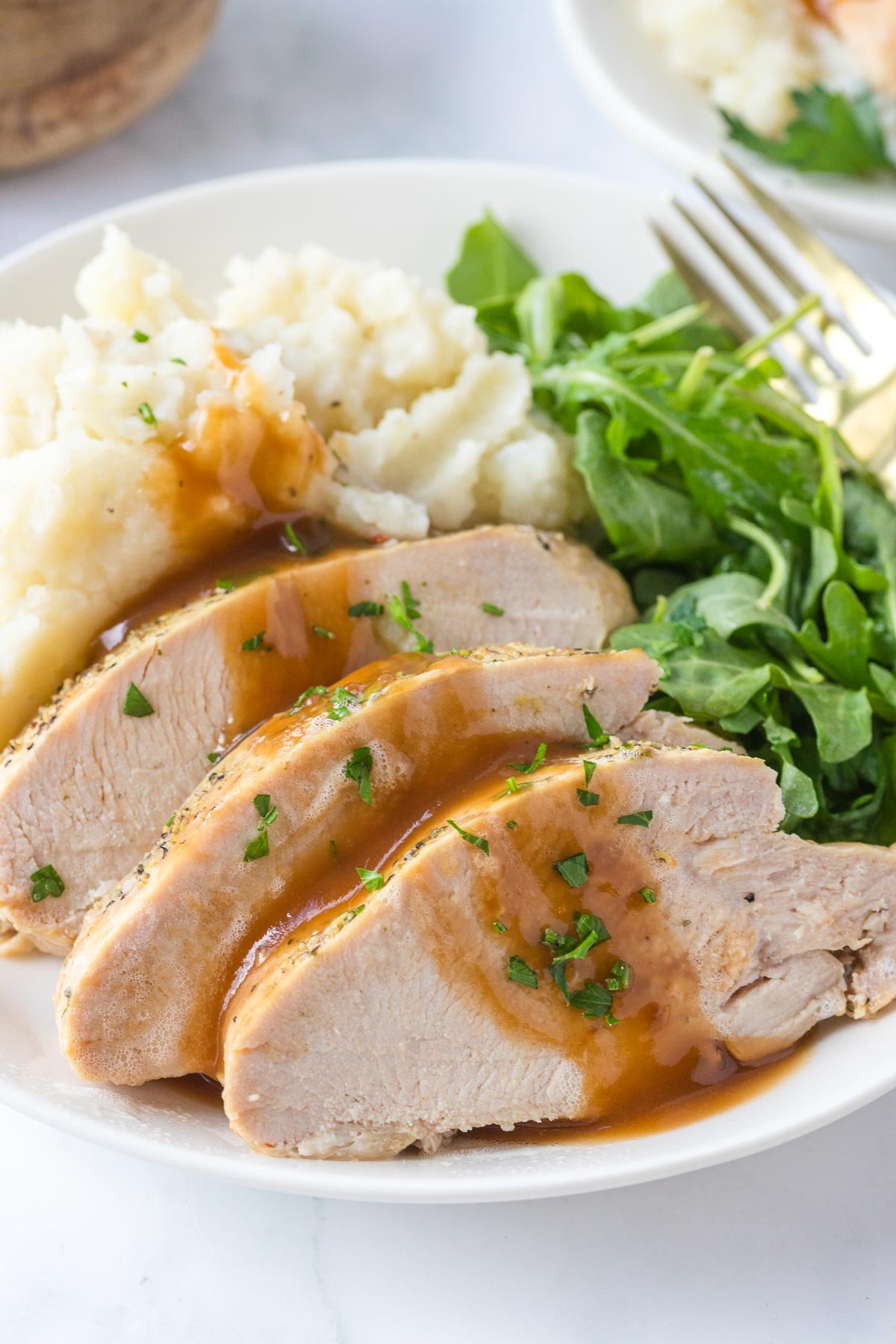 Three slices of slow cooker turkey breast with brown gravy, served with mashed potatoes and a green leafy salad on a white plate with a fork.