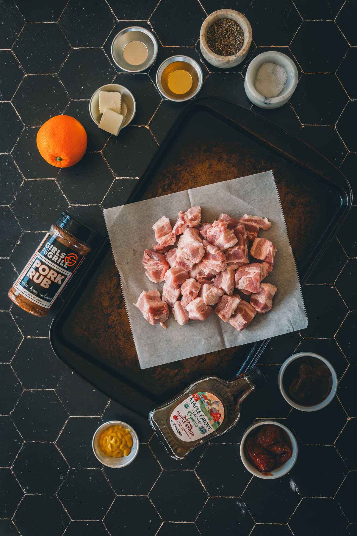 A tray with raw pork belly cubes on parchment paper, surrounded by various seasonings, sauces, an orange, and a bottle of pork rub on a dark tiled surface.