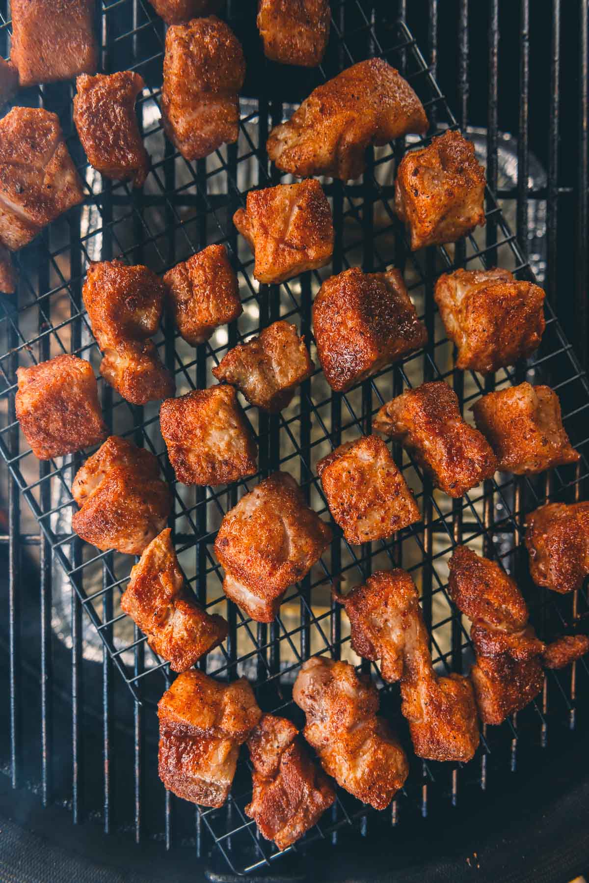Seasoned cubes of smoked pork belly burnt ends are arranged on a wire rack over a grill, cooking gently over indirect heat.