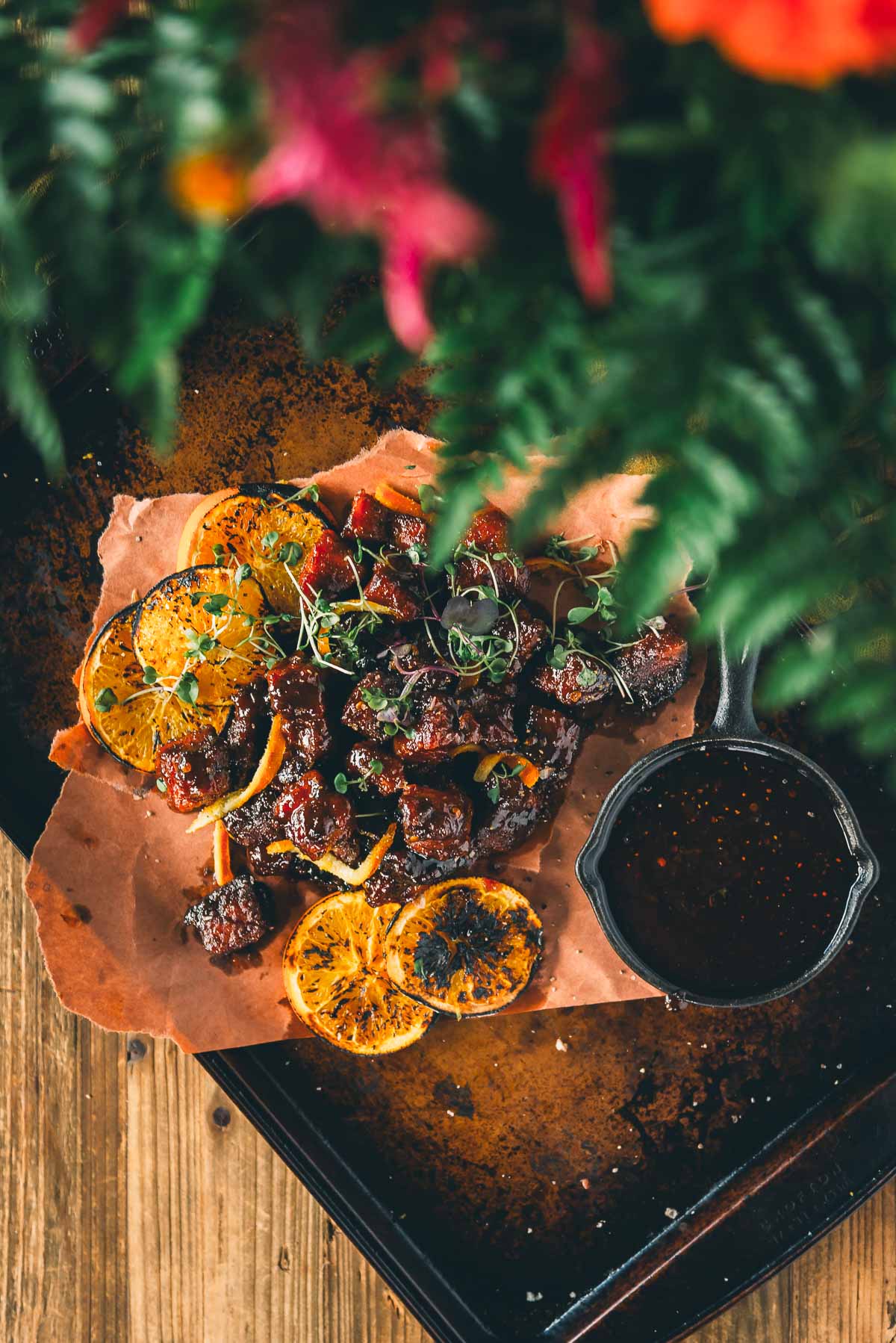 Smoked pork belly burnt ends with charred orange slices on parchment paper, and a dipping sauce; partially obscured by foliage at the top.