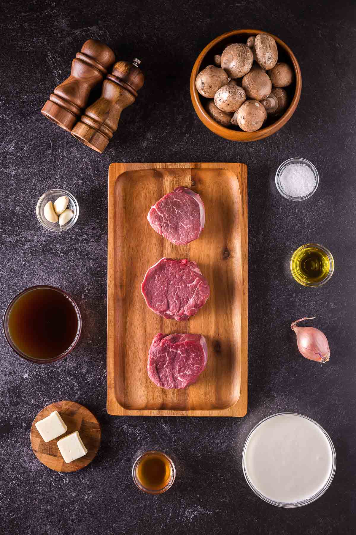 Three raw filet mignon steaks on a wooden tray surrounded by mushrooms, garlic, butter, broth, cream, olive oil, shallot, salt, pepper mills, and a small bowl of sauce on a dark surface.