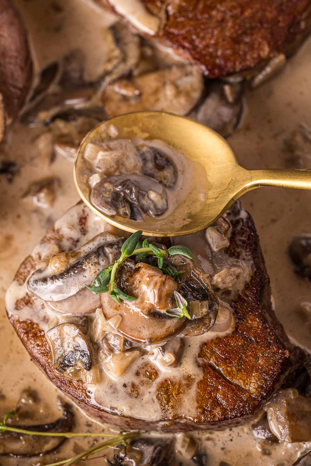 A close-up of a cooked steak topped with creamy mushroom sauce, garnished with fresh herbs, with a gold spoon holding more sauce above it.