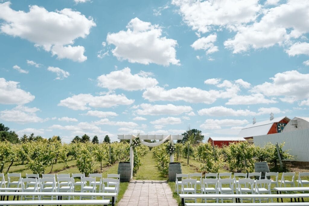 Wine barrels flank a white wooden altar draped in white cloth at an outdoor wedding ceremony set in a winery under blue skies with puffy clouds floating above