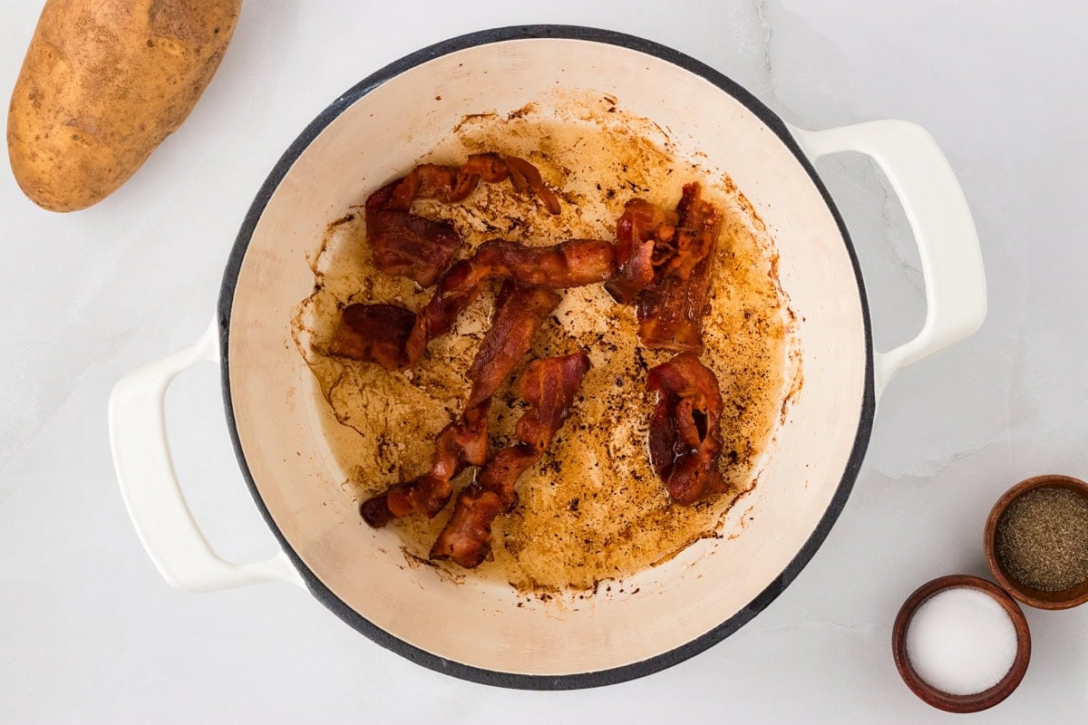 Crispy bacon pieces cooking in a white Dutch oven, showing the rendered bacon fat that will be used to flavor the soup.