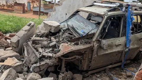 AFP via Getty Images A severely damaged car sitting on rubble. behind it in the background is a patch of grass on the left, a building, and a partial wall on the right