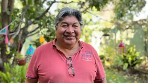 Carlos Alberto Cossio, wearing a pink polo shirt, poses for a photo. He is smiling at the camera while standing in a garden decorated with lights and bunting. His glasses are tucked into his shirt.