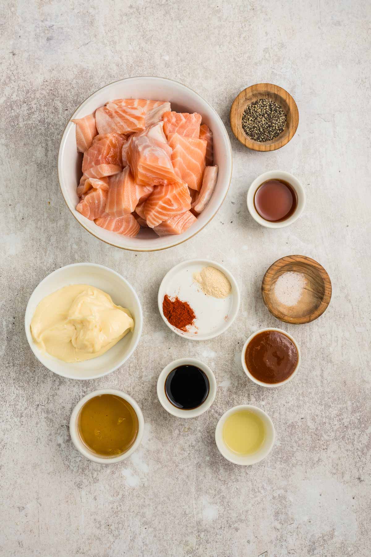 A bowl of raw salmon chunks surrounded by small bowls of mayonnaise, honey, soy sauce, hot sauce, vinegar, spices, and seasonings on a light surface.