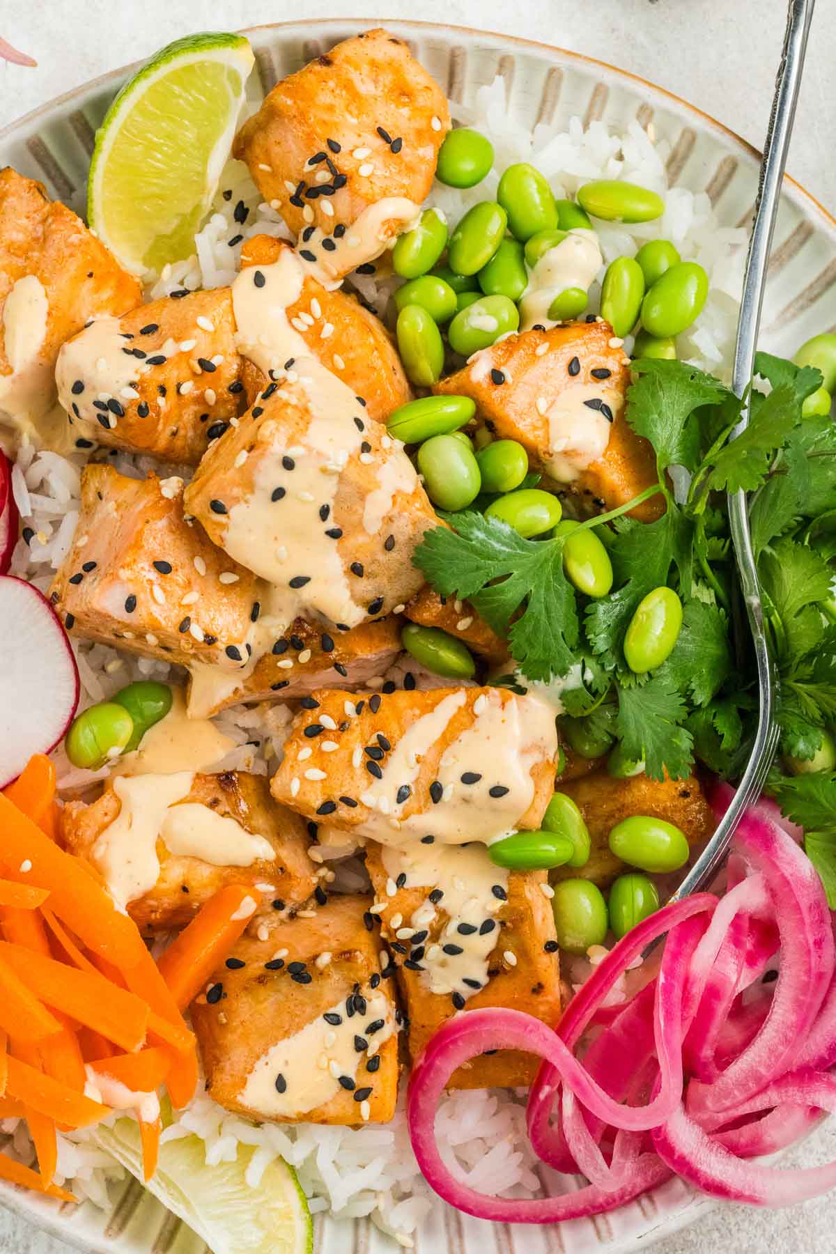 A bowl of rice topped with cooked salmon, edamame, sliced radishes, pickled onions, carrot strips, cilantro, lime, and a creamy sauce, garnished with black sesame seeds.