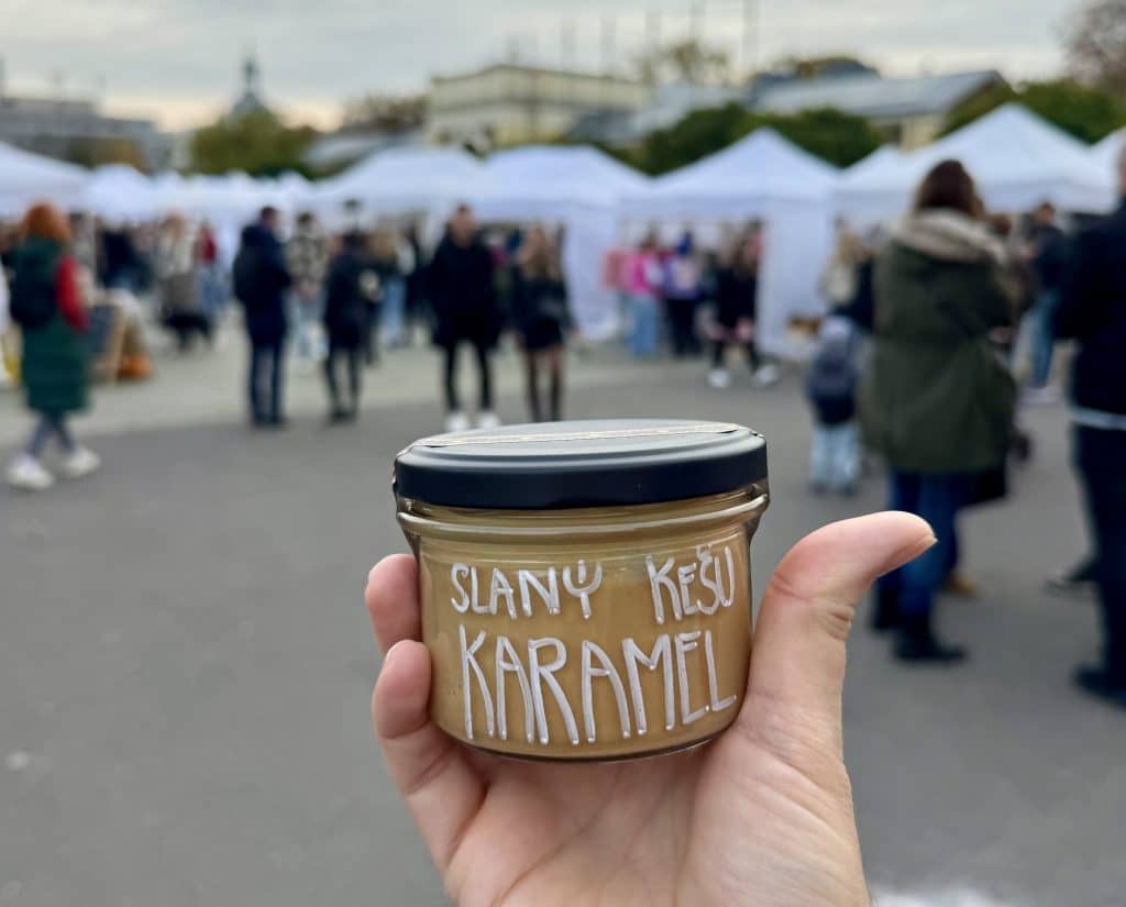 A hand holding a jar of cashew butter with the name written in Czech, with a market of people and tents in the background.