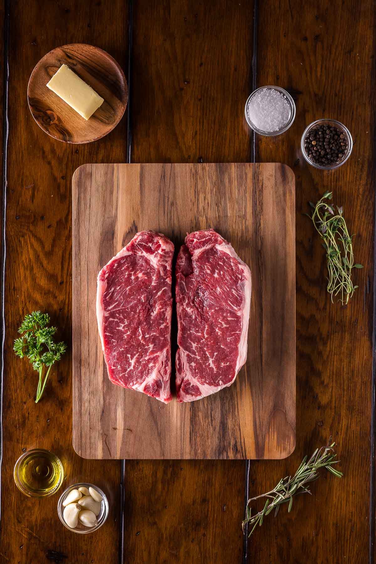 Two raw strip steaks on a wooden cutting board, surrounded by butter, salt, pepper, herbs, garlic, and olive oil on a wooden table.