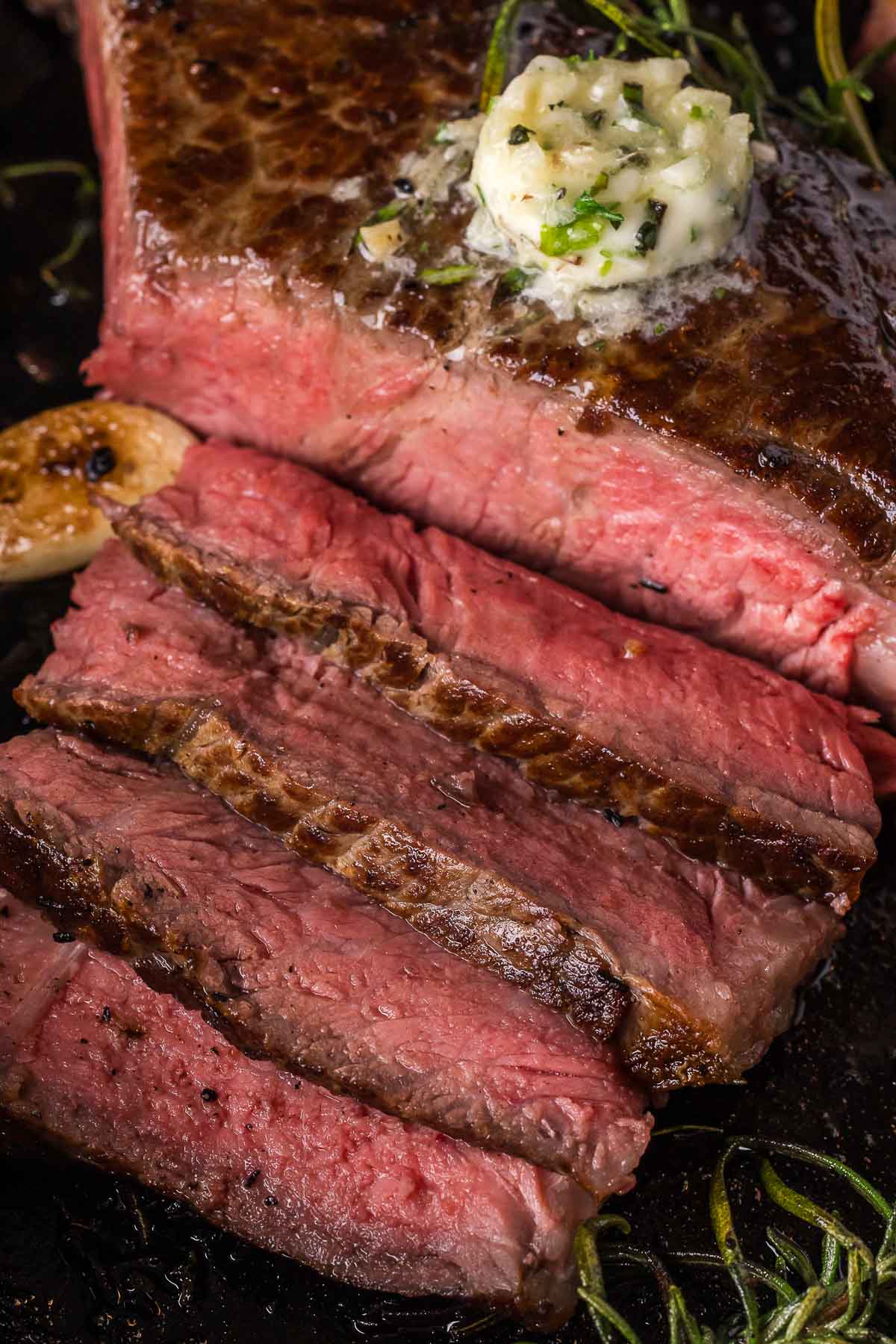 A sliced medium-rare steak on a dark surface, topped with a dollop of garlic butter, with visible rosemary and garlic.
