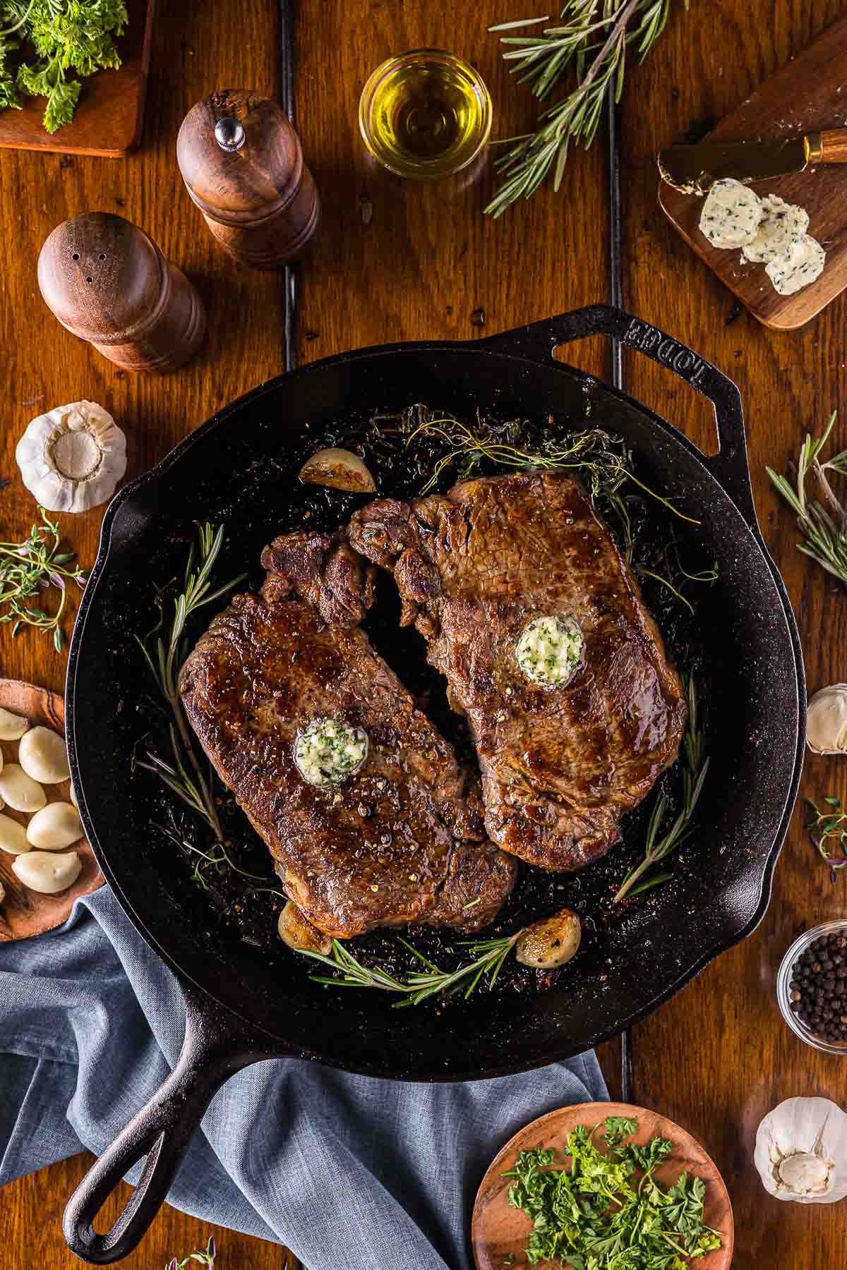 Two cooked steaks with herb garlic butter topped in a cast iron skillet, surrounded by garlic cloves, herbs, pepper, and oil on a wooden table.