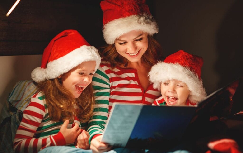 Christmas Eve traditions UK: mum and children in Santa hats reading a Christmas story together