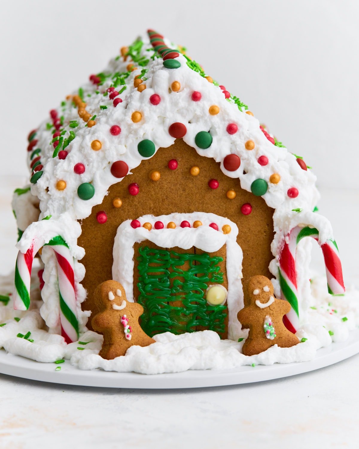 A fully assembled gluten-free gingerbread house decorated with white royal icing, colorful candies, candy canes, and two small gingerbread people by the doorway.