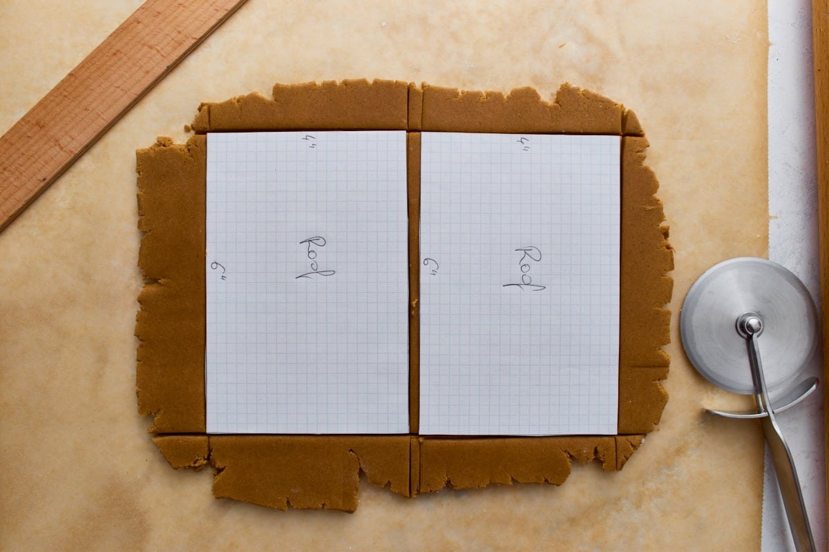 A piece of rolled gingerbread dough with graph-paper roof templates placed on top, ready to be trimmed with a pizza cutter.