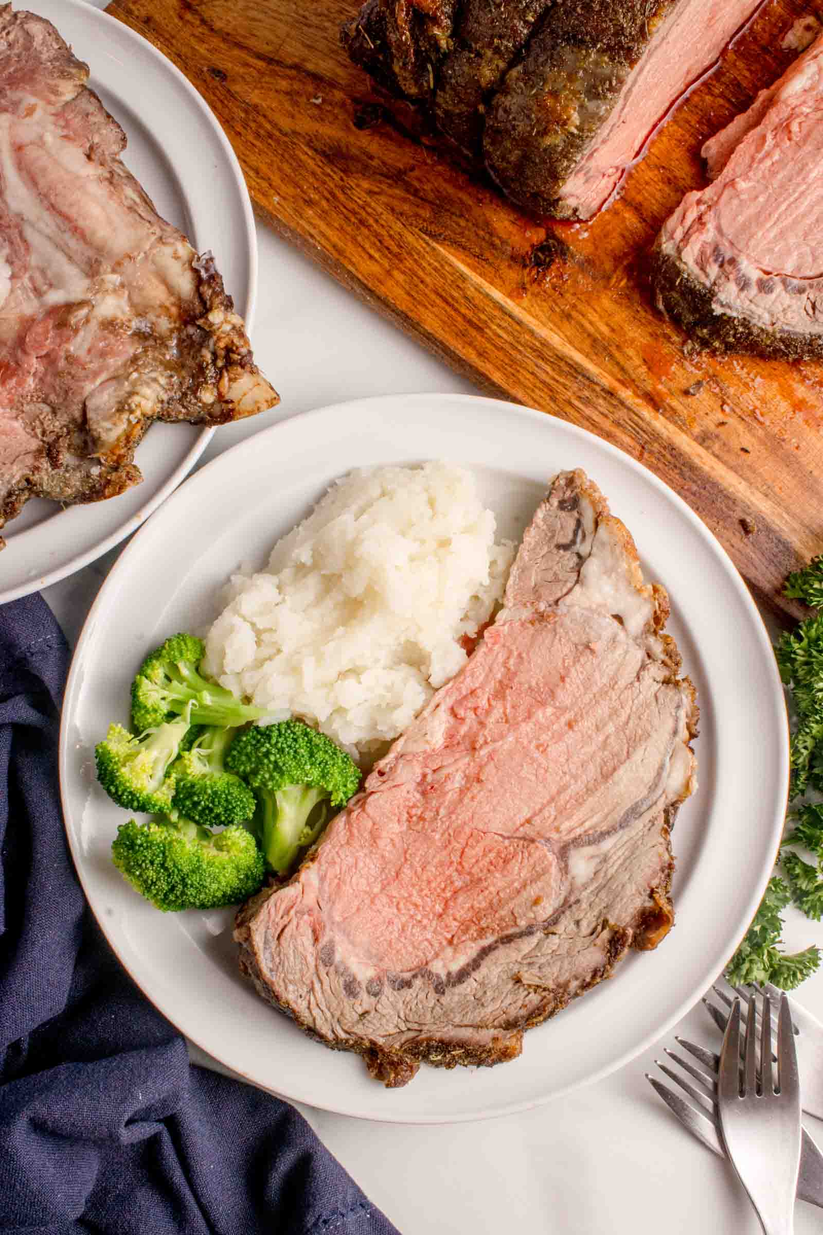 A dinner plate with a slice of prime rib roast, mashed potatoes, and steamed broccoli, next to a cutting board with additional roast beef.