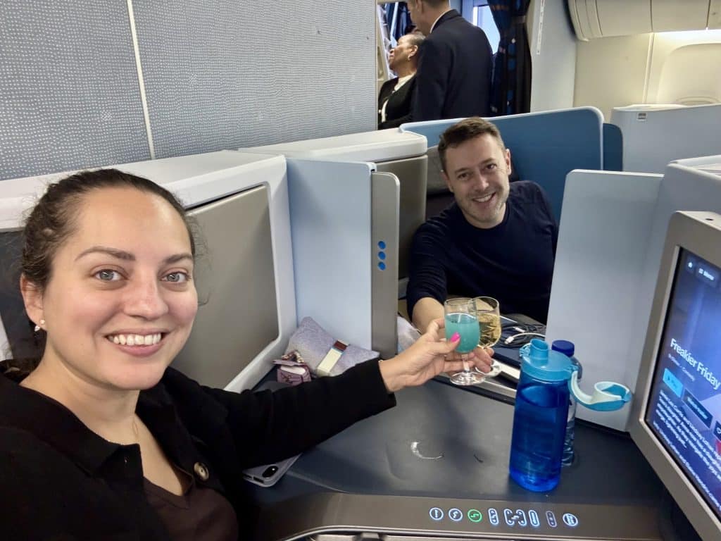 Kate and Charlie taking a selfie in business class, clinking drinks. Kate has a bright blue mocktail and Charlie has a champagne.