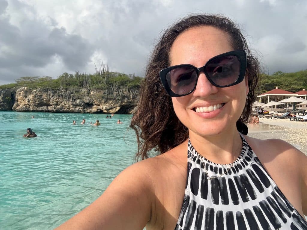 Kate taking a smiling selfie with sunglasses and a black and white bathing suit while standing in bright turquoise water in Curaçao.