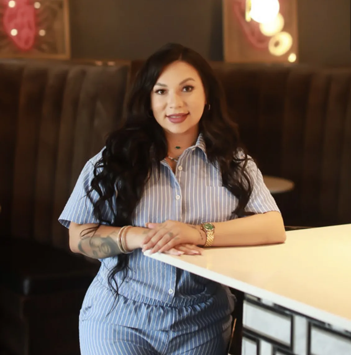 A woman with long, dark hair and a short-sleeve collared shirt stands at the corner of a bar, posing for a photo