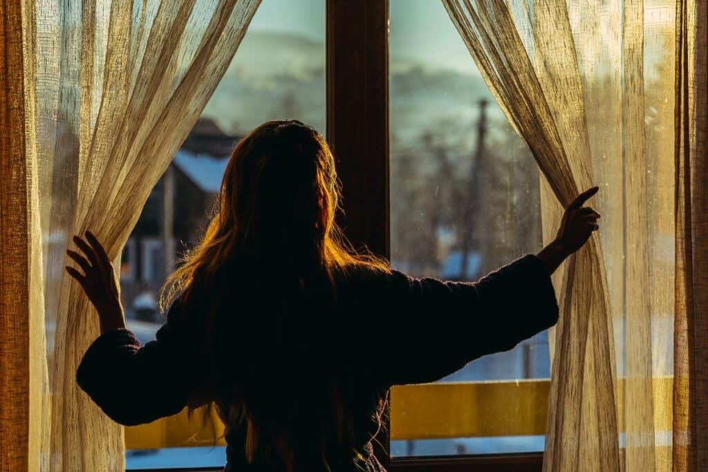 Woman opening curtains at sunrise, winter morning routine for dark mornings