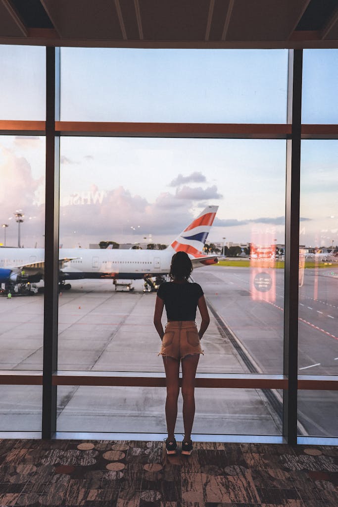 A woman stands at a glass window viewing airplanes at the Airport, waiting to go home after getting sick while travelling solo