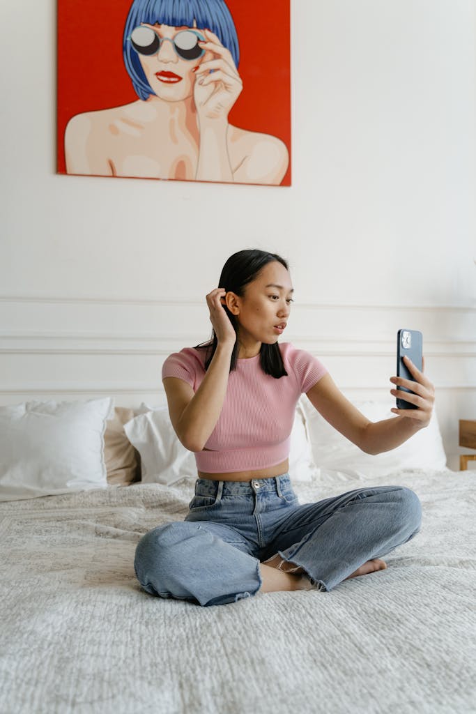A young woman sitting on a bed taking a selfie in a contemporary styled bedroom. Practice makes perfect