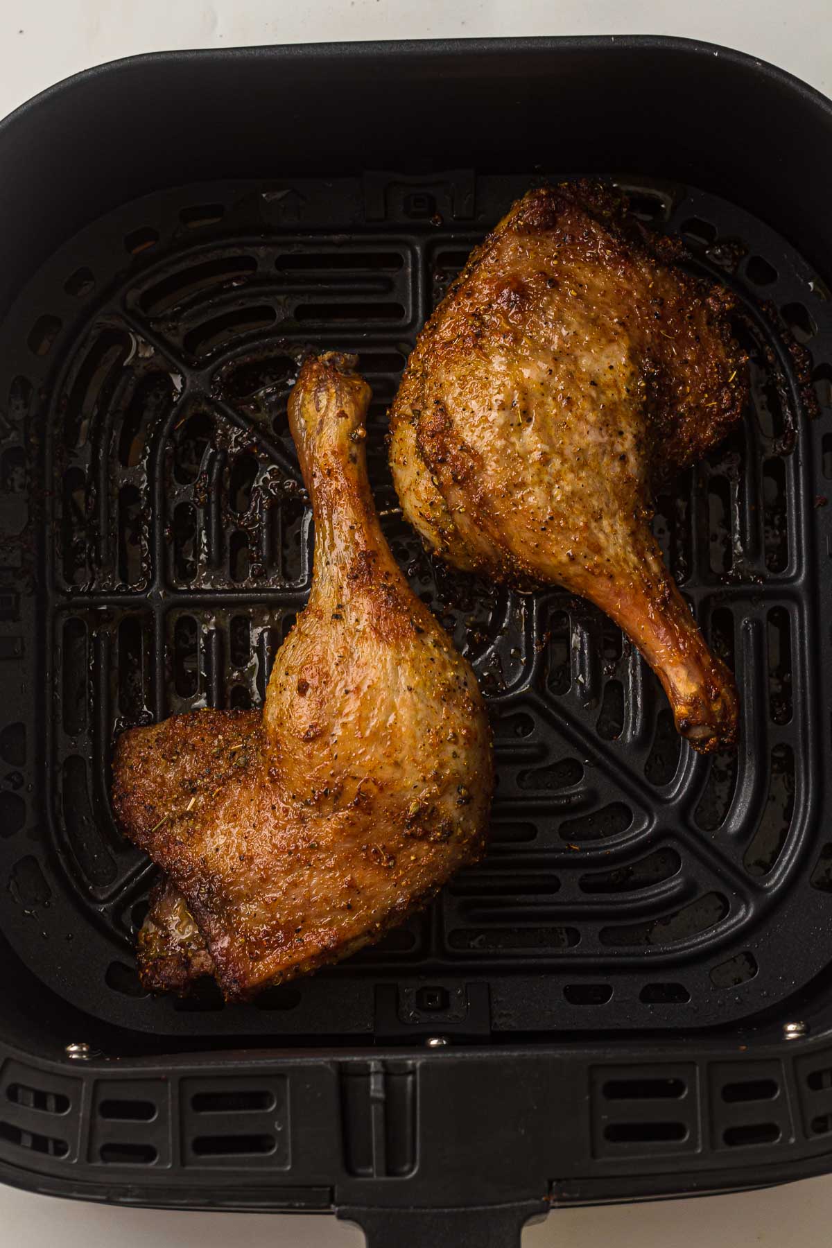 Two cooked duck leg quarters with crispy, browned skin sit in the basket of an air fryer.